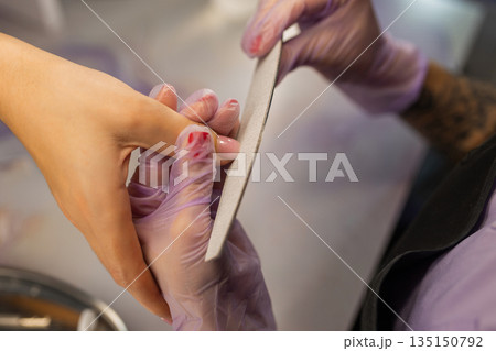 Close up of professional manicure shaping nails with file. Nail technician wearing gloves filing female nails in beauty salon. Nail care, hygiene and cosmetic treatment concept. 135150792