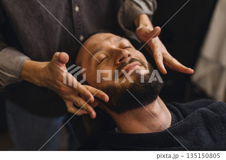 Barber massaging beard and neck of male client after shaving in barbershop. Relaxing grooming service, men self care, comfort and classic barber ritual concept. Barber massaging beard and neck of male client after shaving in barbershop. Relaxing grooming service, men self care, comfort and classic barber ritual concept. 135150805