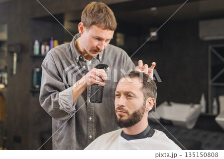 Barber applying hair styling spray to male client during haircut in modern barbershop. Professional grooming service, men hairstyle care, barber work process and lifestyle concept. 135150808