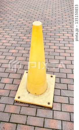 Bright yellow plastic traffic cone standing on a red brick paved sidewalk in a city outdoor area 135150853
