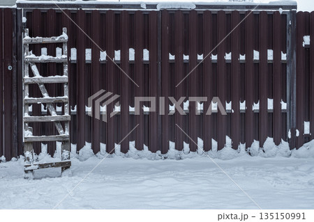 Old wooden ladder leaning against a fence covered with snow. Winter rural background, construction and maintenance concept outdoors. 135150991