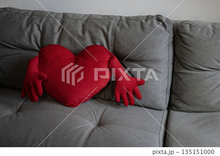 Red heart shaped pillow with arms on gray sofa. Love concept, emotions, relationships, loneliness, Valentine theme. Red heart shaped pillow with arms on gray sofa. Love concept, emotions, relationships, loneliness, Valentine theme. 135151000