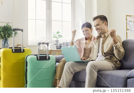 Excited couple booking holiday online at home, cheering with luggage and laptop 135151398