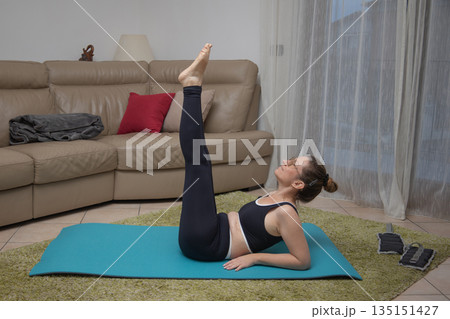 Woman performing a pilates leg raise exercise on a yoga mat in her living room, focusing on core strength and flexibility during a home workout session Woman performing a pilates leg raise exercise on a yoga mat in her living room, focusing on core strength and flexibility during a home workout session 135151427