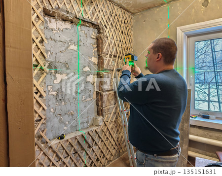 A builder manually uses a high-tech laser level to precisely align an old wall with wood sheathing. Using a laser level, the builder ensures accuracy during a home improvement project. 135151631