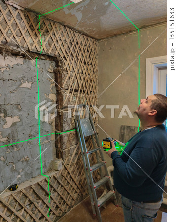 A man with a laser level projects bright green horizontal and vertical lines onto partially destroyed lath and plaster walls. He's taking measurements and planning a home renovation project. A man with a laser level projects bright green horizontal and vertical lines onto partially destroyed lath and plaster walls. He's taking measurements and planning a home renovation project. 135151633