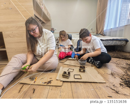 A mother and children sit on the floor and do crafts using wooden sticks, cardboard, scissors, and a glue gun, encouraging creativity and family time. A mother and children are making a craft. 135151636