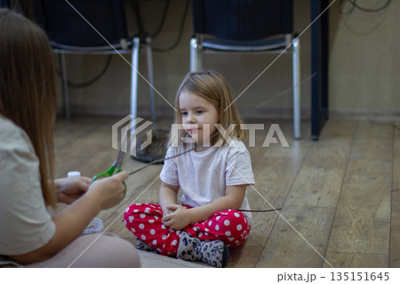 A mother and a small child sit on the floor, making a craft out of a stick with scissors. The family spends time being creative at home, encouraging the child's early development and imagination. A mother and a small child sit on the floor, making a craft out of a stick with scissors. The family spends time being creative at home, encouraging the child's early development and imagination. 135151645
