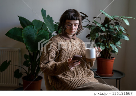 Teen girl sitting on chair with cup of tea, reading news, scrolling photos, messaging in smartphone Teen girl sitting on chair with cup of tea, reading news, scrolling photos, messaging in smartphone 135151667