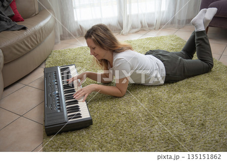 Woman lying on green shag rug in living room, casually playing an electronic keyboard, barefoot in white socks, enjoying a relaxed music practice and cozy creative downtime at home Woman lying on green shag rug in living room, casually playing an electronic keyboard, barefoot in white socks, enjoying a relaxed music practice and cozy creative downtime at home 135151862