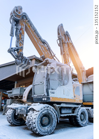 Scenic view frozen hydraulic excavators standing outdoors winter conditions covered snow ice industrial building alpine area. Cold weather impacts machinery operation hydraulic systems reliability 135152152