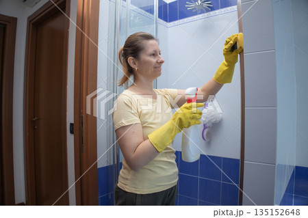 Woman wearing yellow rubber gloves and holding a spray bottle, diligently cleaning bathroom shower tiles to maintain a hygienic and sparkling home environment Woman wearing yellow rubber gloves and holding a spray bottle, diligently cleaning bathroom shower tiles to maintain a hygienic and sparkling home environment 135152648