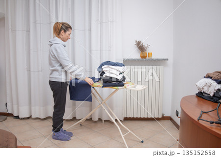 Woman folding clean clothes and organizing a pile of ironed garments on an ironing board inside a bright room at home, performing daily domestic chores Woman folding clean clothes and organizing a pile of ironed garments on an ironing board inside a bright room at home, performing daily domestic chores 135152658