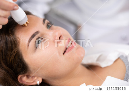 Unrecognizable beautician doctor doing RF lifting procedure for flawless woman face laying in beauty salon. Hardware cosmetology Unrecognizable beautician doctor doing RF lifting procedure for flawless woman face laying in beauty salon. Hardware cosmetology 135155916