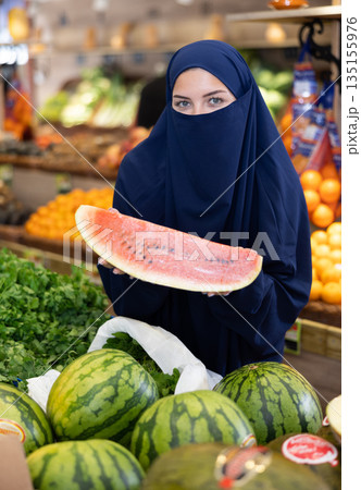 Girl in veil carefully chooses fresh watermelon in supermarket Girl in veil carefully chooses fresh watermelon in supermarket 135155976