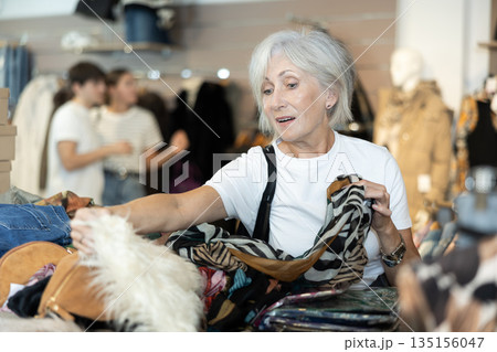 Senior woman buyer carefully sort through various wardrobe items presented in window of second-hand store 135156047