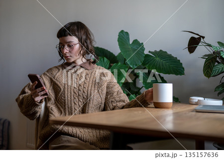 Thoughtful girl student sits at desk with cup, reading news in cellphone procrastination of homework 135157636