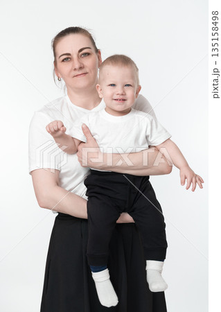 Middle aged Caucasian mother holding 1 year old boy's child in her arms and hugging darling son. Mommy and toddler boy looking at camera. Female parent and kid wearing white t-shirts and black bottoms 135158498