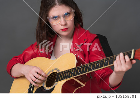 Close-up view of female guitarist strumming strings of her classical acoustic guitar while looking directly at camera. Stylish musician is young Caucasian woman wearing glasses and red leather jacket Close-up view of female guitarist strumming strings of her classical acoustic guitar while looking directly at camera. Stylish musician is young Caucasian woman wearing glasses and red leather jacket 135158501