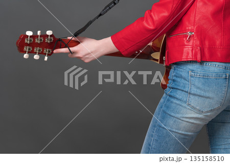 Side view of unrecognizable woman guitarist strumming her Spanish six string acoustic guitar. Crop view of midsection of female musician dressed in crimson leather jacket and blue jeans 135158510