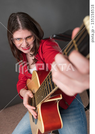 Woman playing guitar in lighthearted manner, appears to be deriving enjoyment from activity. She is doing so with smiling. Young female exudes passion and is attired in crimson leather jacket, glasses Woman playing guitar in lighthearted manner, appears to be deriving enjoyment from activity. She is doing so with smiling. Young female exudes passion and is attired in crimson leather jacket, glasses 135158518