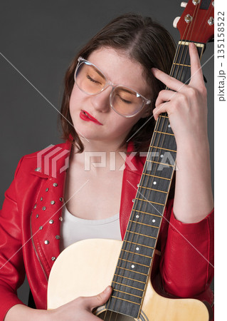 Lady with guitar gently nestles musical instrument against her bosom. A young female musician wearing glasses and red leather jacket 135158522