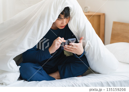 布団にくるまりスマートフォンを操作している若い男性 ライフスタイルイメージ 布団にくるまりスマートフォンを操作している若い男性 ライフスタイルイメージ 135159208