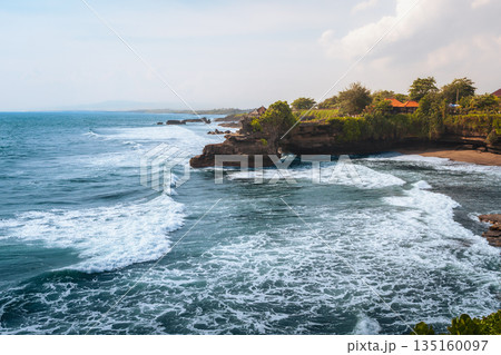 Tanah Lot temple Bali ocean waves crashing rock, iconic Hindu shrine on cliff amid turquoise sea lush greenery blue sky exotic tropical Indonesian coastal paradise serene landscape travel destination 135160097