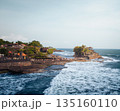 Tanah Lot temple Bali ocean waves crashing rock, iconic Hindu shrine on offshore cliff amid turquoise sea lush greenery orange flags blue sky exotic tropical Indonesian coastal paradise landscape 135160110