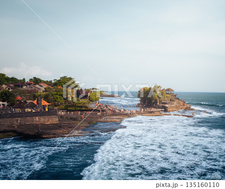 Tanah Lot temple Bali ocean waves crashing rock, iconic Hindu shrine on offshore cliff amid turquoise sea lush greenery orange flags blue sky exotic tropical Indonesian coastal paradise landscape 135160110
