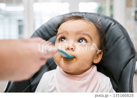 Adorable, sweet and cute baby eating puree for lunch, dinner or snack in her high chair at home. Child development, food and girl infant kid enjoying meal with spoon for growth and wellness in house. 135160828