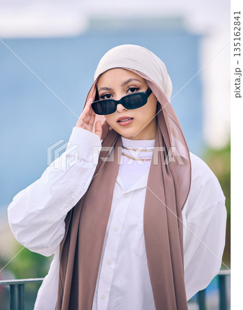 Portrait, fashion or sunglasses with a muslim woman in dubai wearing a cap and scarf for contemporary style. Islamic, faith and hijab with a trendy young arab person posing outside in modern eyewear Portrait, fashion or sunglasses with a muslim woman in dubai wearing a cap and scarf for contemporary style. Islamic, faith and hijab with a trendy young arab person posing outside in modern eyewear 135162841