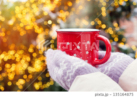 Female hands in warm knitted gloves holding a red mug with a hot drink  against the lights of a Christmas tree in winter 135164875