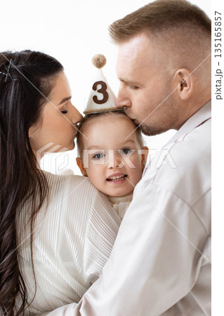 A loving family celebrates a child's third birthday with kisses. The parents embrace their daughter, who wears a party hat. 135165857