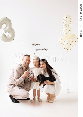 A family of three celebrates a little girl's third birthday with a cake and a dachshund dog in a bright, minimalist setting. 135165896
