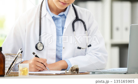 Planning, doctor writing on a clipboard and with laptop at a desk in a modern office with lens flare. Healthcare, medical research and female nurse or surgeon with stethoscope write notes at work 135165905