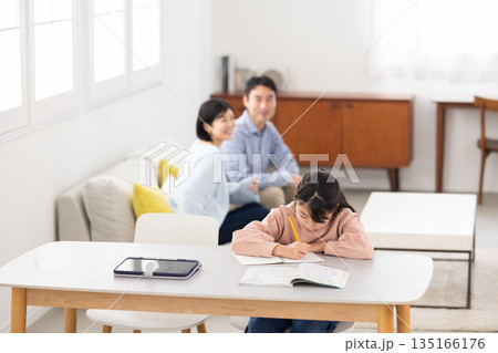 両親が見守る中リビングのテーブルで勉強する小学生の女の子 135166176