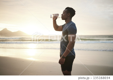 Fitness, beach and black man drinking water at sunset on running break, exercise or mockup. Drink, liquid and African male athlete with bottle for nutrition, hydration or health, wellness and ocean. 135168034