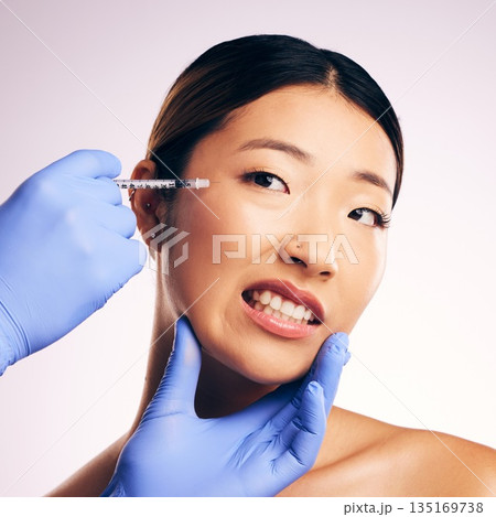 Face injection, skincare and Asian woman in pain in studio isolated on a white background. Cosmetics, syringe and female model with collagen filler, dermatology and prp facelift in plastic surgery. 135169738