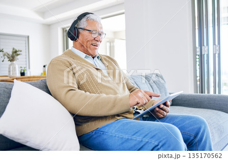Old man laughing, relax with tablet and headphones at home, retirement and technology, streaming comedy online. Surfing internet, watching funny video and elderly male person sitting on couch 135170562
