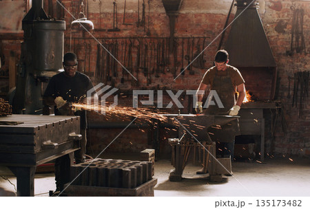 Hammer, anvil and sparks with men working in a forge for metal work manufacturing or production. Industry, welding and trade with a young male blacksmith in a workshop, plant or industrial foundry 135173482
