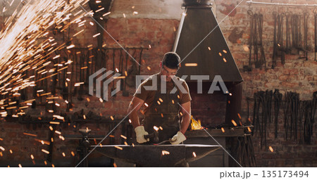 Hammer, anvil and sparks with a man at work in a forge for metal work manufacturing or production. Industry, welding and trade with a male blacksmith working in a factory, plant or industrial foundry Hammer, anvil and sparks with a man at work in a forge for metal work manufacturing or production. Industry, welding and trade with a male blacksmith working in a factory, plant or industrial foundry 135173494