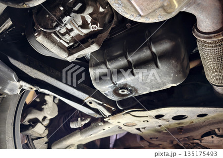 A person inspects the oil pan and engine components beneath a car in a garage. There is visible dirt and grease on the parts from use A person inspects the oil pan and engine components beneath a car in a garage. There is visible dirt and grease on the parts from use 135175493