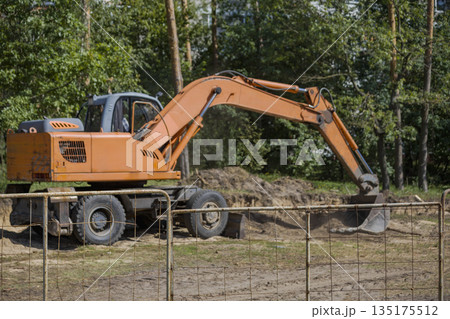 A large excavator digs into the ground at a construction site surrounded by trees during day. Workers may be nearby managing the project 135175512