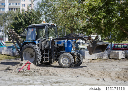 A tractor is digging on a construction site in the city during daylight. Nearby, there are parked cars and stacked materials for the project 135175516