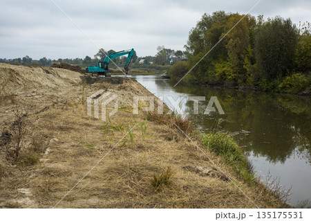 A machine is digging near the riverbank while trees and bushes line the opposite shore. The sky is overcast and the water is still A machine is digging near the riverbank while trees and bushes line the opposite shore. The sky is overcast and the water is still 135175531
