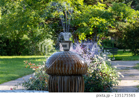 Water fountain features a creative design with splashing water, surrounded by lush greenery. 135175565