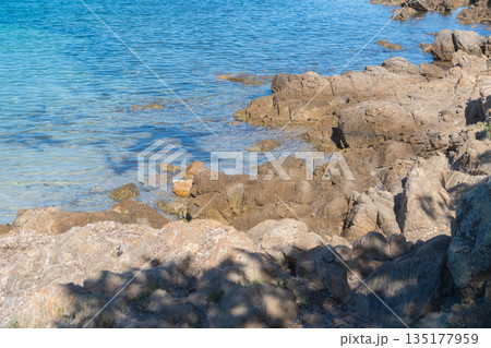 Shaded rocks meet clear shallow water along a quiet coastline. Sunlit blue tones blend with rugged textures, creating a peaceful seaside mood perfect for travel, nature detail, and coastal landscape Shaded rocks meet clear shallow water along a quiet coastline. Sunlit blue tones blend with rugged textures, creating a peaceful seaside mood perfect for travel, nature detail, and coastal landscape 135177959