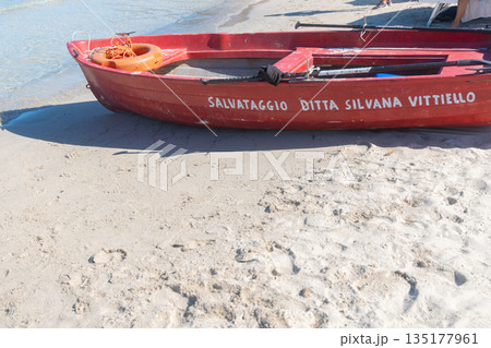 A red wooden boat rests on soft sand near the calm shoreline. Gentle waves and clear daylight create a bright coastal setting with strong color contrast, ideal for travel, nautical, and beach 135177961