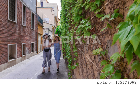 Two attractive laughing women in panamas walking between the buildings and having fun Two attractive laughing women in panamas walking between the buildings and having fun 135178968